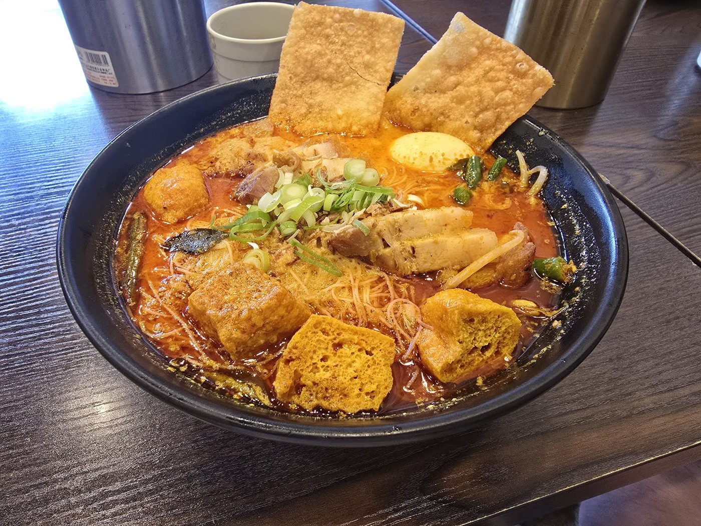 Curry laksa in a black bowl: tofu puffs, rice vermicelli, long beans, half an egg and two big blistered crisp sheets standing on top, in a red curry broth